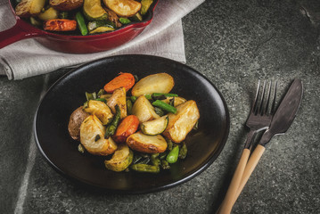 Plate and Skillet with  fried seasonal autumn vegetables (zucchini, potatoes, carrots, beans), on black stone table copy space