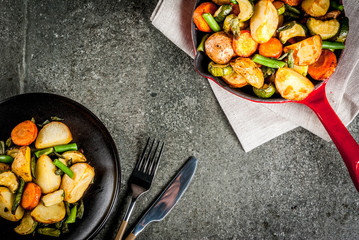 Plate and Skillet with  fried seasonal autumn vegetables (zucchini, potatoes, carrots, beans), on black stone table copy space top view