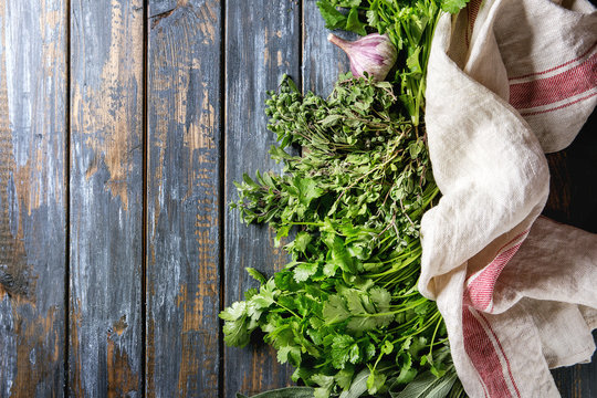 Variety of fresh organic herbs coriander, sage, oregano with garlic and textile kitchen towel over old wooden plank background. Top view with copy space. Food background