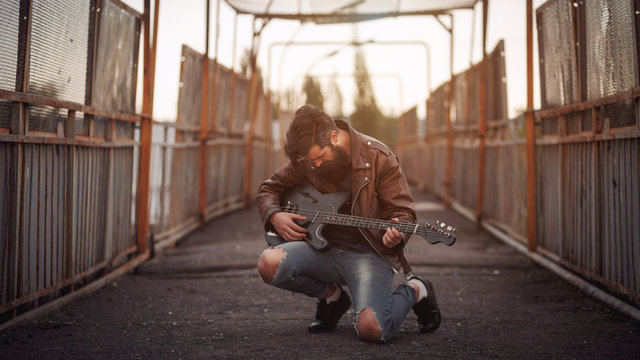 Stylishly dressed handsome man with a long beard, brown leather jacket and light jeans playing guitar on the bridge