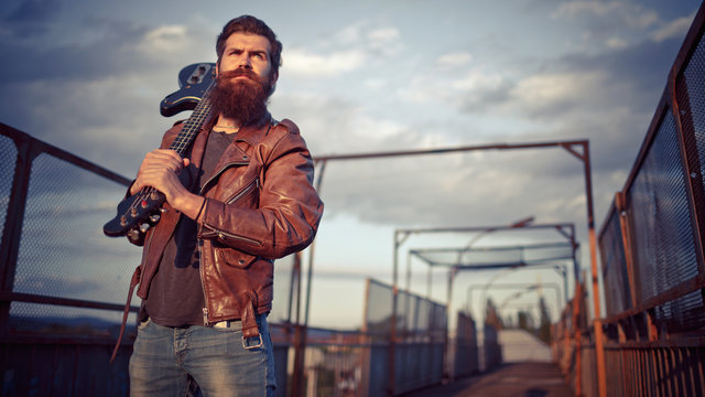 Stylishly dressed handsome man with a long beard, brown leather jacket and light jeans holding a guitar and standing on a bridge