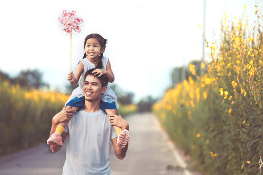 Cute asian little girl playing with wind turbine and riding on father's shoulders in the flower garden in vintage color tone. Father and daughter having fun to play together.