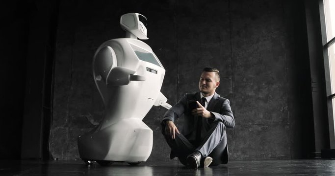 businessman uses a smartphone while sitting on the floor next to a robot. Modern Robotic Technologies. Humanoid autonomous robot.