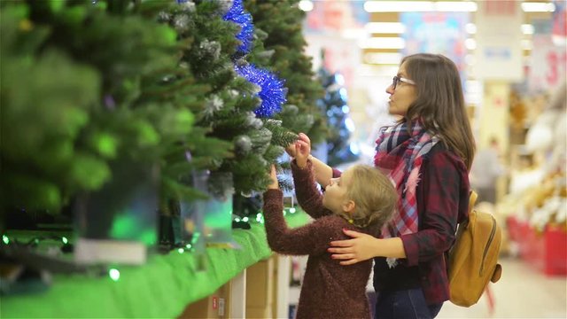 Mother and Child Choosing Christmas tree in the Supermarket. Young Beautiful Mom and Daughter Buys Xmas-tree with Decorations near the Supermarket Shelf, Merry Christmas and Happy New Year