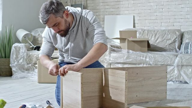 PAN of concentrated bearded man sitting on floor of new flat and putting screws into wooden shelf