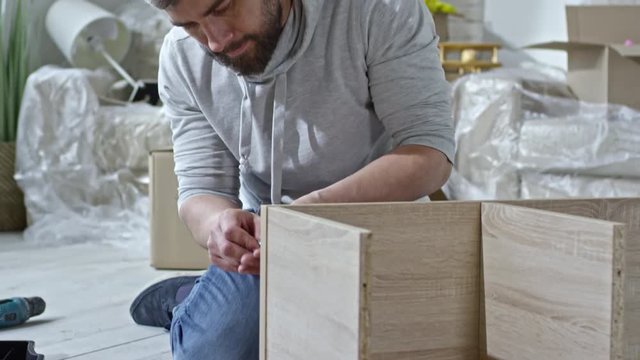 Tilt up of bearded man in hoodie sitting on floor of new flat and assembling wooden shelf; cardboard boxes and sofa covered in plastic wrap standing in background
