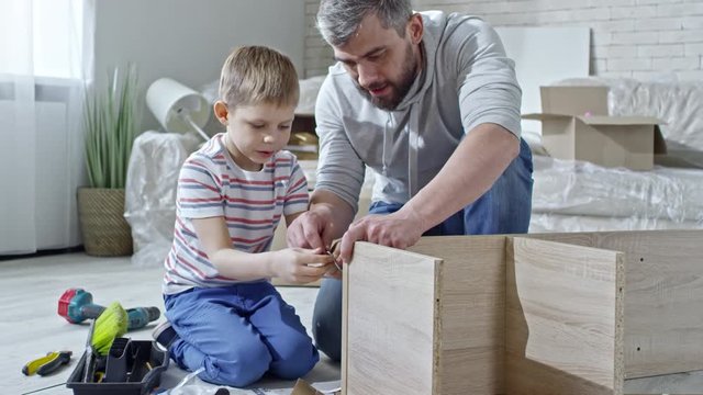 PAN of cheerful father with beard and cute little boy sitting on floor of new apartment and building wooden shelf together