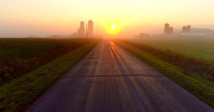 Sunrise over foggy rural agricultural landscape with farms and silos, aerial view.
