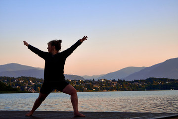Mature woman in black clothes doing yoga on a wood jetty on Faaker See in Austria, early in the morning at sunrise