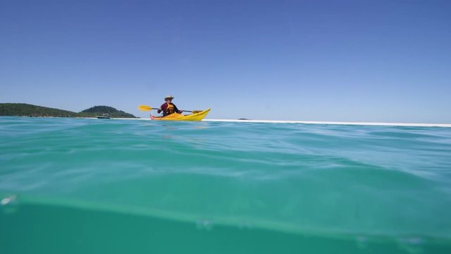 An over - under shot of a beach that shows a yellow kayak and a man on the ocean surface. The background shows a white sand shore and a green mountain. The footage was shot in slow motion.