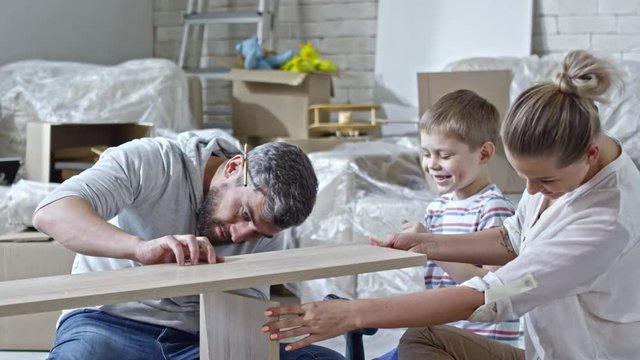 Slow motion of beautiful young woman holding wooden pieces while little boy giving tools and helping bearded father assembling shelf