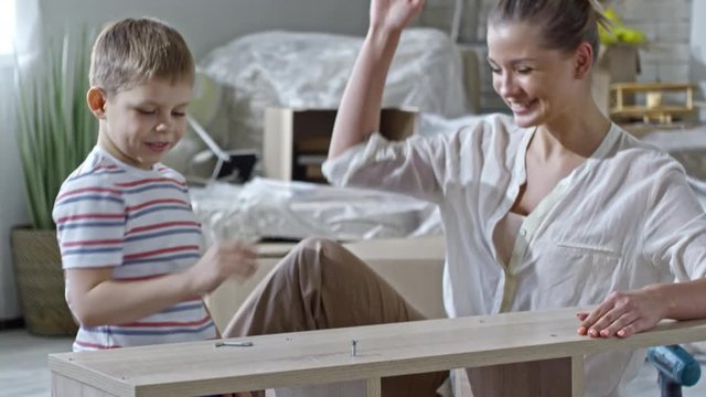 Tilt up of beautiful mother high-fiving son helping her to assemble wooden shelf in new apartment