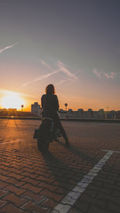 Outdoor lifestyle portrait of sexy biker girl wearing leather jacket sits on a modern motorcycle, sunset over city in the background