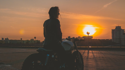 Naklejka premium Outdoor lifestyle portrait of sexy biker girl wearing leather jacket sits on a modern motorcycle, sunset over city in the background