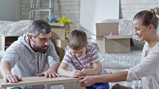 PAN of little boy with blond hair helping bearded father and beautiful mother assembling wooden shelf, then high-fiving dad
