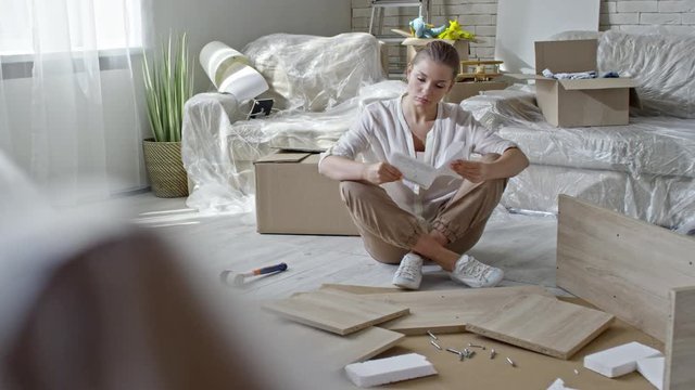 PAN of tired woman sitting on floor of new house and reading assembling manual, then looking at disassembled wooden furniture and thinking; sofa in plastic wrap and unpacked boxes in background