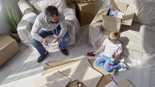 Top view of bearded man in hoodie sitting in armchair covered with plastic wrap and talking with little son holding drill and looking at disassembled furniture