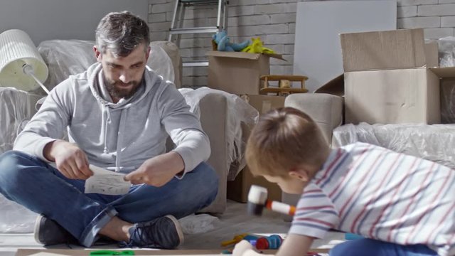 Zoom out shot of little boy playing with toy hummer and disassembled furniture while bearded man sitting and reading manual