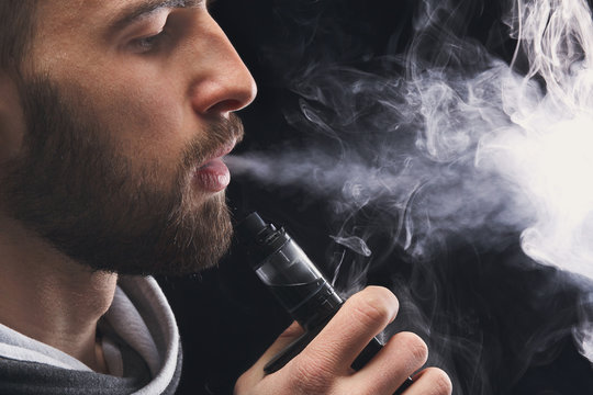Young man vaping e-cigarette with smoke on black