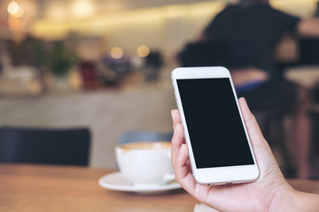 Mockup image of hands holding white mobile phone with blank black screen with coffee cups on wooden table in restaurant
