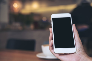 Mockup image of hands holding white mobile phone with blank black screen with coffee cups on wooden table in restaurant
