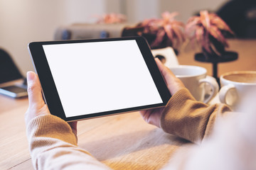 Mockup image of hands holding black tablet pc with blank white screen with coffee cups on wooden table in modern cafe