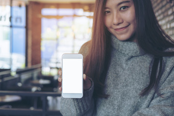 Mockup image of an Asian beautiful woman holding and showing white mobile phone with blank  screen in cafe with brick wall background