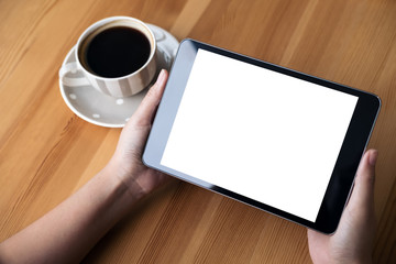 Mockup image of business woman's hands holding black tablet pc with blank white screen and coffee cup on wooden table in cafe background