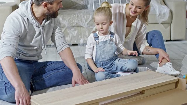 Tilt up of cute little girl in denim overalls sitting on floor of new house with bearded father and beautiful mother and helping them open cardboard box with wooden furniture, then looking at manual