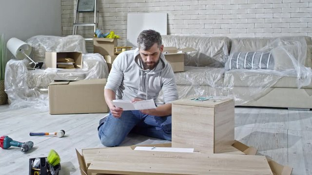 PAN of concentrated man with beard sitting on floor of new apartment and reading assembling manual for furniture, then looking at wood screws; sofa covered with plastic wrap in background