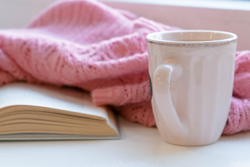 Cozy home still life: cup of hot coffee and opened book with warm plaid on windowsill against snow landscape outside. Winter holidays, Christmas time concept, free copy space
