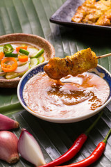 A hand having pork satay dipping peanut sauce decorated with cucumber sauce, onion, chili on banana leaf.