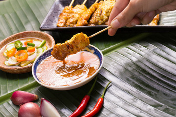 A hand having pork satay dipping peanut sauce decorated with cucumber sauce, onion, chili on banana leaf.