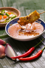 A hand having pork satay dipping peanut sauce decorated with cucumber sauce, onion, chili on banana leaf.