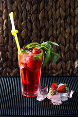 Red cocktail lemonade with strawberries and ice cubes on red napkin with strawberries and mint leaves on black background in dark bar table