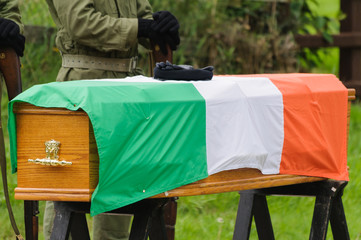 Fototapeta na wymiar IRA paramilitary gloves and beret on a coffin with an Irish Tricolour flag. IRA man in background.