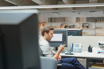 Young businessman checking messages on mobile phone at desk in busy office after hours