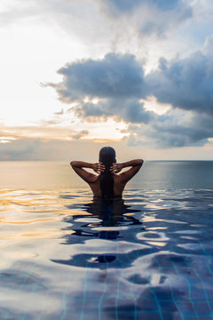 Woman at the edge of swimming pool with sea horizon and sky in background during sunset.