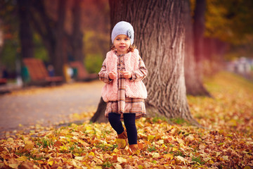 cute fashionable baby girl walking in autumn park among fallen leaves