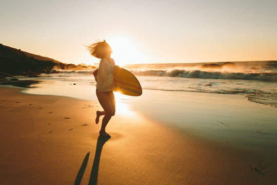 Young woman surfer with surfboard running on beach at sunset