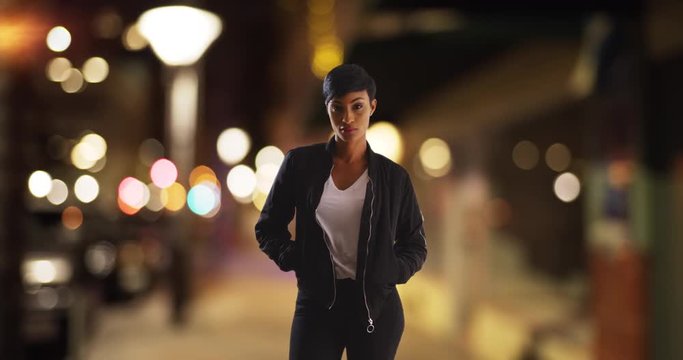 Black female in stylish jacket posing with hands in pockets on city street in evening. Portrait of African American woman standing in urban setting, looking at camera with serious expression 