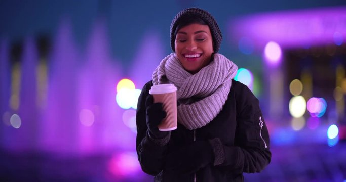 Portrait of lovely black female wrapped in scarf, standing by fountain side in evening laughing and smiling. Cute woman in her 20s holding coffee cup outdoors on cold fall night 
