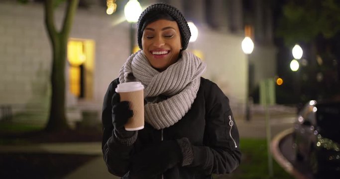 Cute African American woman wrapped in scarf and beanie standing outside college library, laughing and smiling. Portrait of black female holding coffee cup outdoors on cold night 