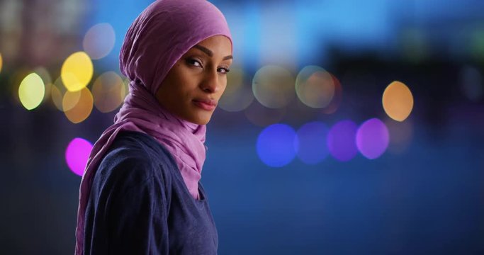 Portrait of black female in purple headscarf looking at camera with serious expression. Muslim woman standing in urban setting at night with pretty blue bokeh lights 