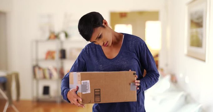 Beautiful black female shaking cardboard box excitedly, guessing what's inside. Attractive woman examining package in living room, smiling joyfully 