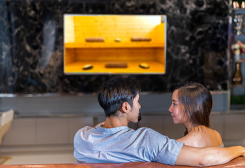 Young couple together watching tv with sauna spa in resort program and sitting sofa in living room at home happy romantic resting relax for honeymoon in holidays, back view