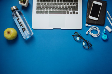 Creative Office table for healthy person with laptop, pencils, water, apple, phone, glasses, headphones on a blue background. Top view, flat lay, space for text