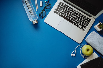 Workspace for healthy person with laptop, pencils, water, apple, phone, glasses, headphones on a blue background. Top view, flat lay, space for text