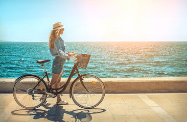 Blonde woman in summer hat with her bicycle walking coastline by