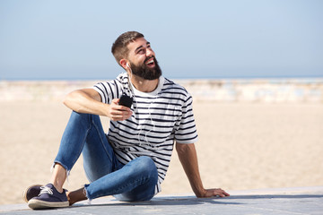Fototapeta na wymiar Full length happy man in earphones sitting on beach with mobile phone